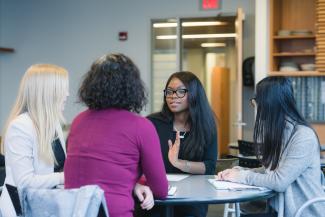 Four people seated around a table engaged in a collaborative discussion in a professional setting, with one person gesturing while speaking.