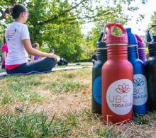 Colourful, reusable water bottles from the the UBC Yoga fund in the foreground, A group meditates in the background on the grass.